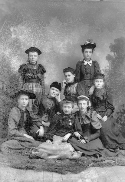 A group of eight girls, six sitting and two standing, pose for a studio portrait in front of a painted backdrop.