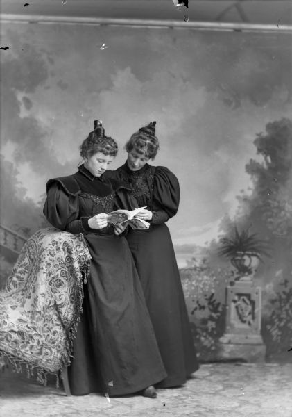 A studio portrait of two women standing near a chair and reading from the same book in front of a painted backdrop. They both wear similar black dresses and hairstyles.