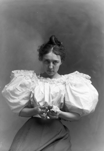 A studio portrait of a young woman in a puffy-sleeved blouse with a skirt holding an unidentified object, possibly a handkerchief.