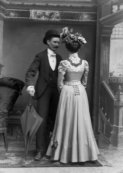 A well-dressed couple stands for a studio portrait in front of a painted backdrop. The man holds an umbrella and wears a hat, suit jacket, vest, bow tie, and trousers. The woman wears a long dress and hat and stands with her back to the camera.