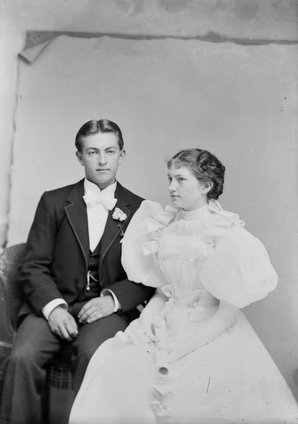 Studio portrait of a man wearing a suit jacket with a flower in the lapel, vest with watch fob, bow tie, and trousers. A woman sits next to him and is wearing a white dress with puffy sleeves. The woman holds a rolled piece of paper, probably a diploma.