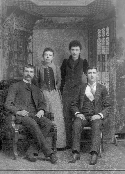 A group of four individuals, two men and two women pose for a studio portrait in front of a painted backdrop. The men wear suit jackets, vests, and ties, and sit while the women in long dresses stand behind them.