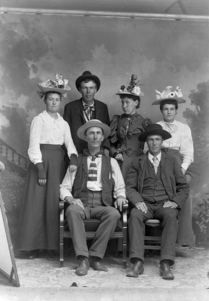 Studio portrait in front of a painted backdrop of two men sitting in front of three women and a man who are standing. They are all wearing hats. The men are wearing suit jackets, vests, and ties, and the women are wearing long dresses or skirts.