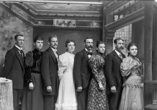 Three-quarter length studio group portrait in front of a painted backdrop of young people in a European American family posed standing. The woman wear dresses with puffed sleeves, the men wear suits and neckties. Identified as probably the Oderboltz family. From left to right: Pat Oderboltz, Julia Oderboltz, Charles Oderboltz, Mary Oderboltz, Frank Oderboltz, Anna Oderboltz, George Oderboltz, and Emma Oderboltz.