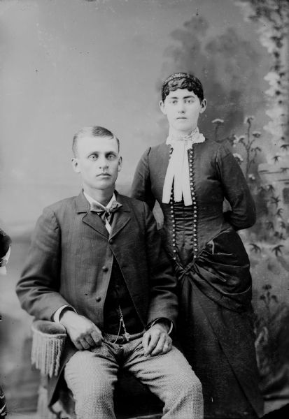 A studio portrait of a man and woman posed in front of a painted backdrop. The man wears a suit jacket, vest with watch fob, and trousers and sits in a chair in front of the woman who is standing behind him, resting her hand on the back of the chair.