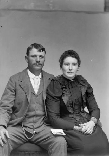 A seated man and woman pose for a studio portrait. The man wears a suit jacket, vest with watch fob, and trousers and he has his arm around the woman. A piece of paper rests on the man's knee, possibly a photograph.