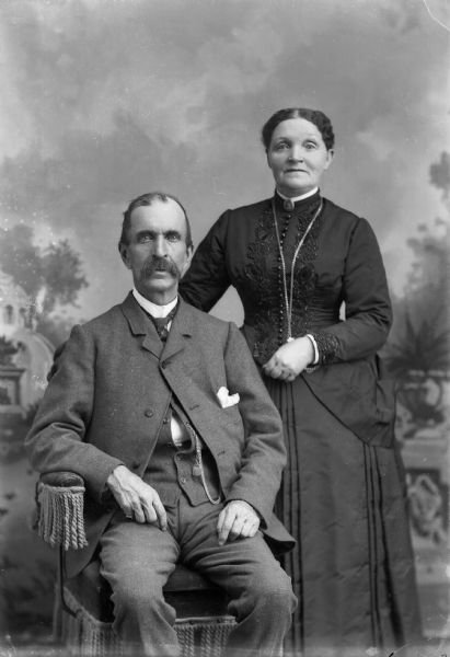 A man wearing a suit jacket, vest with watch fob, and trousers, sits in a chair as a woman wearing a dress stands next to him. They are posing for a studio portrait in front of a painted backdrop.