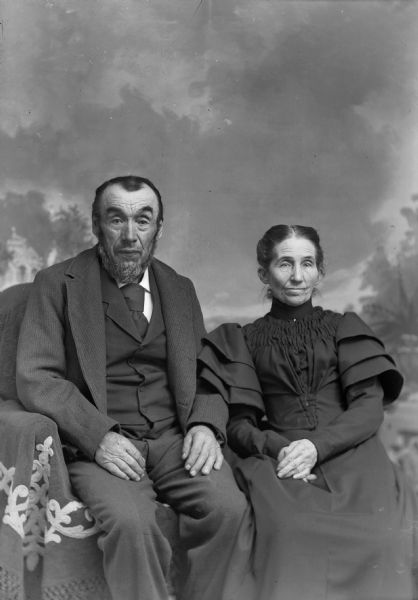 An elderly man and woman sit for a studio portrait in front of a painted backdrop. The man wears a suit jackets, vest, and trousers, and the woman wears a dress with ruffled sleeves.