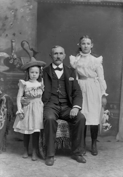 A seated elderly man sits between two standing girls in front of a painted backdrop. The man wears a suit jacket, vest with watch fob, tie, and trousers, and has his arm around the younger girl, while the older girl stands with her hand behind the old man's chair.