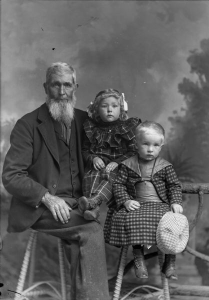 A studio portrait of a seated elderly man with two young children in front of a painted backdrop. The elderly man wears a suit jacket, vest, and trousers, and holds a young girl wearing a dress on his lap. A young boy in a suit sits on a stool holding a hat.