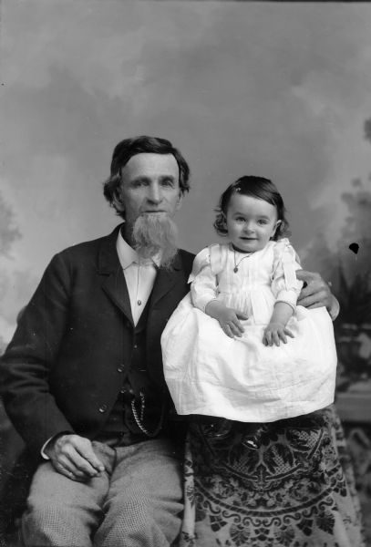 A studio portrait of an elderly man sitting with a small girl in front of a painted backdrop. The man is wearing a suit jacket, vest with watch fob, and trousers. The girl wears a dress and sits on an elevated table and the elderly man steadies her with his arm.