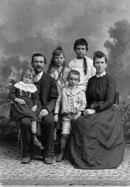A family with four children poses for a studio portrait in front of a painted backdrop. The two parents are seated while two older children stand behind them. A young boy in a shirt and short pants stands between the seated parents and a young girl in a dress sits on her father's lap. The woman wears a long dress, the man a suit jacket, vest, tie, and trousers.