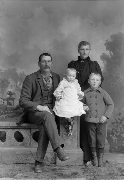 A studio portrait of a couple with two children posing in front of a painted backdrop. A man wearing a suit jacket, vest, tie, and trousers sits on a studio prop of a stone wall with a young child in a white dress. A woman in a long dress, and a boy on a jacket and short pants, stand nearby.