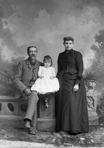 A studio portrait of a man and small girl in a white dress sitting on a studio prop of a stone wall. The man wears a suit jacket, vest, and trousers. A woman in a long dress is standing next to them in front of a painted backdrop.