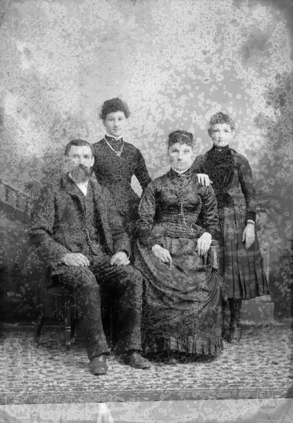 A studio portrait in front of a painted backdrop of a seated man and woman. Two girls stand behind the seated adults, one resting her hand on the seated woman's shoulder. The women are wearing long dresses, the man a suit jacket, vest with watch fob, and trousers.