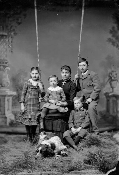 A studio portrait in front of a painted backdrop of a woman sitting on a swing, holding a young child and surrounded by three others. A dog rests at her feet. The woman and girls wear dresses, the boys wear suit jackets and trousers.