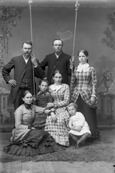 A group of men, women, and children gather around a swing  in front of a painted backdrop and pose for a studio portrait. Two men stand behind the group wearing suit jackets and trousers. A woman stands on the right, two women sit on the swing, and another sits on the floor. A young child sits on a block and rests against one of the women on the swing. All the women are wearing long dresses.