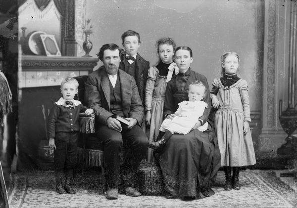 A studio portrait of a seated man, and a woman holding a child, surrounded by two girls and two boys posing in front of a painted backdrop. The man and boys are wearing suit jackets and trousers. The woman and girls are wearing long dresses.