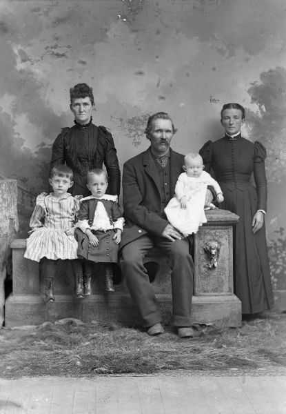 A studio portrait  in front of a painted backdrop of a man with a moustache sitting on a prop stone wall with three children, one on his lap. Two women in dresses stand in the background.