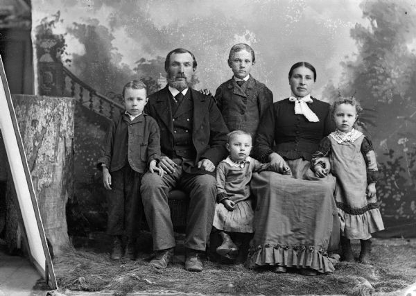 A studio portrait of a seated man and woman surrounded by two boys and two girls in front of a painted backdrop. On the left can be seen the edge of the backdrop, a prop stump, and a canvas covered frame. The men and boys are wearing suit jackets and ties, the woman and girls are wearing long dresses.