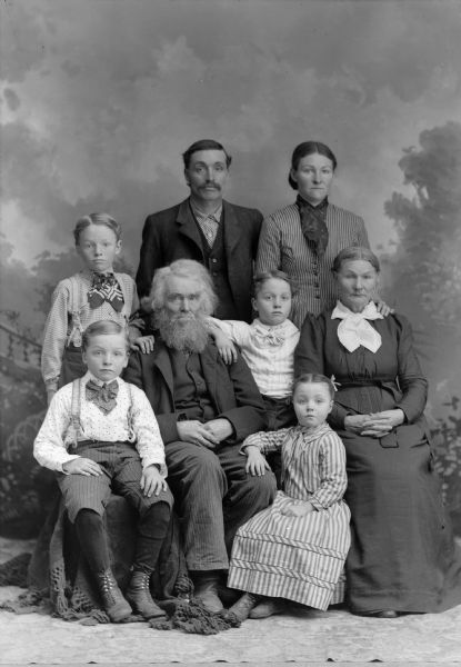 An elderly couple sit surrounded by two other adults standing behind them, and four children, two boys and two girls. They are posing in front of a painted backdrop. The elderly man has a full beard and moustache, and white hair.