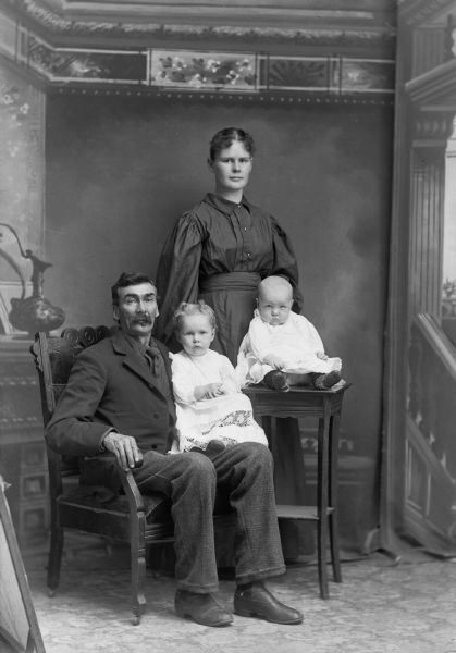 A studio portrait of a seated man in a suit jacket, vest, and trousers holding a young girl on his lap. A woman in a dress is standing behind a baby on a table. They are posed in front of a painted backdrop.