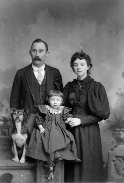 A studio portrait of a standing man and woman, and a small girl and dog sitting on a prop stone wall in front of a painted backdrop. The woman and girl are wearing dresses, the man is wearing a suit jacket, vest, tie, and trousers.