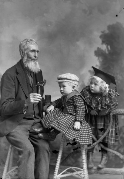 A studio portrait, in front of a painted backdrop, of an elderly man with a beard sitting and holding a stuffed bird to attract the attention of two children. The girl on the fence wears a dress and a mortarboard. The other child, perhaps a boy, sits on a stool wearing a hat and matching jacket and skirt.