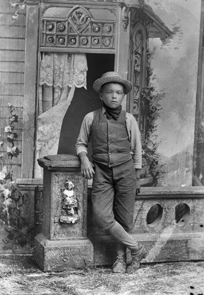 A boy leans against a prop stone wall for a studio portrait in front of a painted backdrop. He is wearing a vest, handkerchief around his neck, a hat, and short pants.