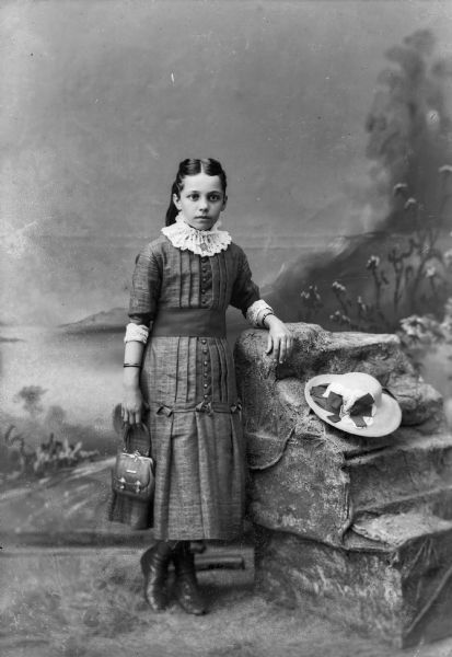 A well-dressed girl leans against a studio prop rock for a portrait in front of a painted backdrop. She is wearing a dress, bracelets, and is holding a purse in her right hand. A hat sits on the rock.