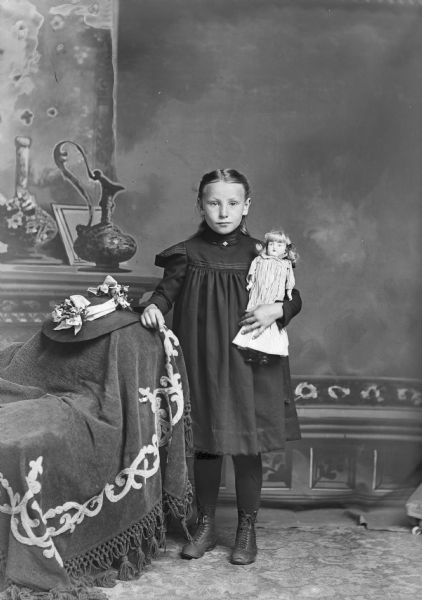 A studio portrait of a young girl wearing a dress and holding a doll in front of a painted backdrop. The girl stands and rests her arm on a stuffed chair where a hat also sits.