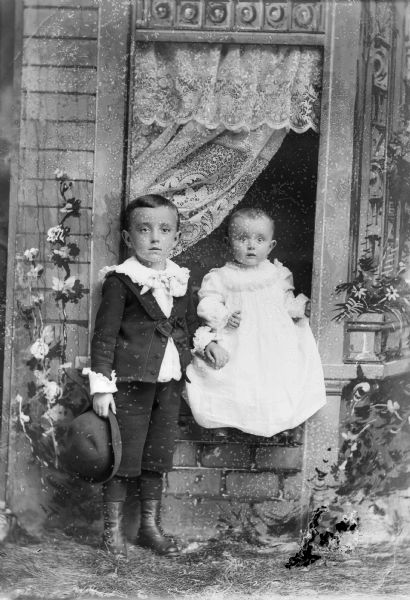 A studio portrait of a small boy holding a hat standing next to a baby girl in a dress seated in a windowsill in front of a painted backdrop. The boy wears a suit jacket, shirt with ruffled collar, and short pants.