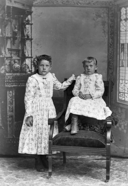 Two girls wearing dresses pose for a studio portrait in front of a painted backdrop. One girl sits on a pillow on a chair while the other stands next to it.