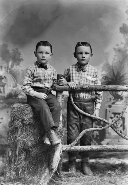 Two young boys pose for a studio portrait in front of a painted backdrop. One stands behind a wooden fence while the other sits on a bale of hay. They both wear plaid shirts, bow ties, and short pants.