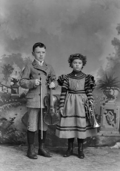 A boy and girl stand for a studio portrait with musical instruments in front of a painted backdrop. The boy wears a matching suit jacket and short pants and holds a violin. The girl wears a dress with ruffled sleeves and holds a cornet.