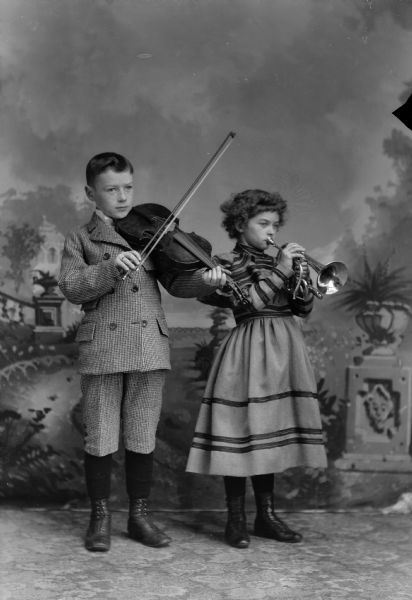A young boy and girl posing standing for a studio portrait, playing musical instruments in front of a painted backdrop. The boy is wearing a matching suit jacket and short pants and is playing a violin. The girl is wearing a dress with ruffled sleeves and is playing a cornet.
