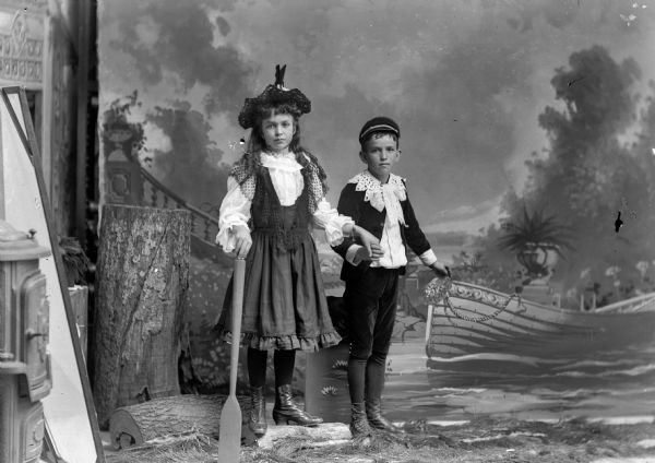 A studio portrait of a girl and a boy in a river scene painted on a backdrop. The girl wears a dress and hat and rests a hand on an oar and holds the boy's hand with the other. The young boy wears a hat, awide-collared shirt over a suit jacket and short pants, and rests his hand on a boat painted on a board.