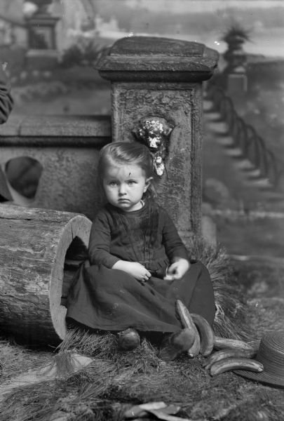 A studio portrait of a small girl with a bell in her hands, sitting by a stone railing, a hollowed-out log, with bananas and a hat scattered near her feet. There is a painted backdrop in the background.