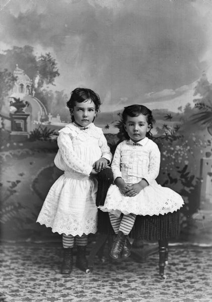 Two small girls pose for a studio portrait in front of a painted backdrop. One is seated while the other stands.