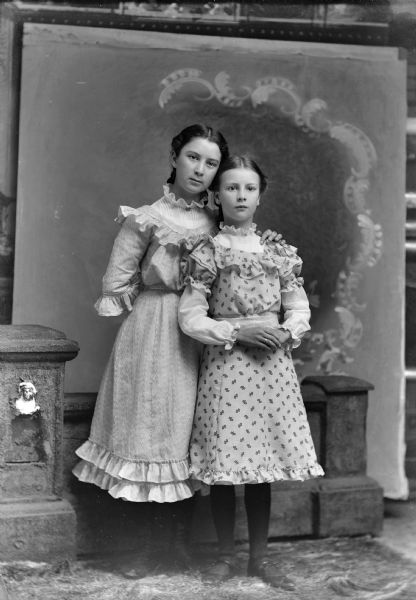 A studio portrait of two standing girls in front of a painted backdrop. One stands with her arm around the other.
