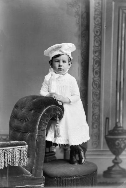 A studio portrait of a small child wearing a hat, dress, and boots, standing on a stool and leaning on a stuffed chair in front of a painted backdrop.