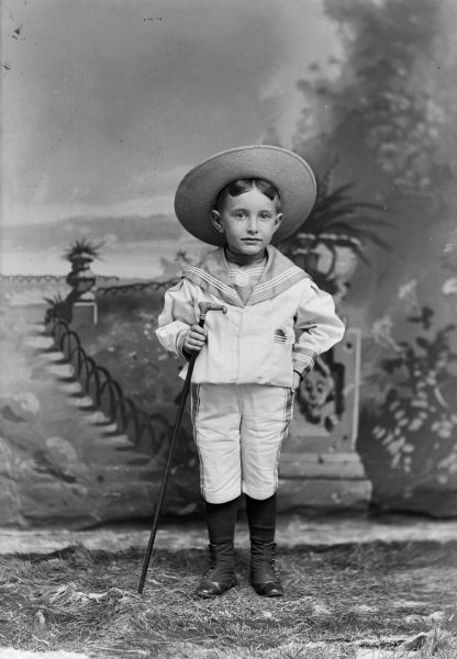 A studio portrait of a small boy, posed in front of a painted backdrop, wearing a hat and holding a cane.