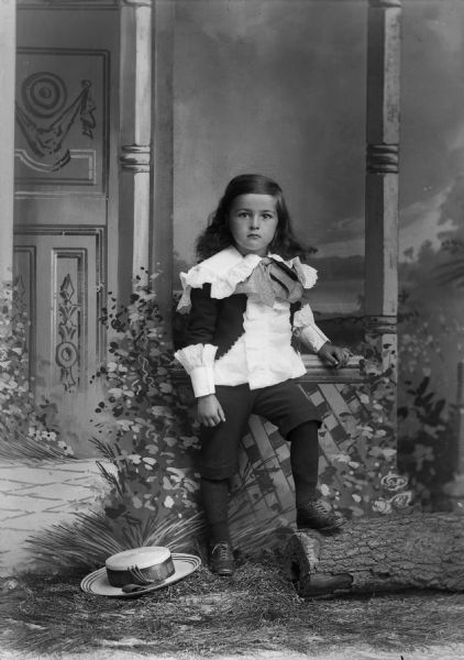 A young child stands for a studio portrait in front of a painted backdrop. The child has long hair and wears a ruffled shirt with short pants. A hat is on the ground nearby.