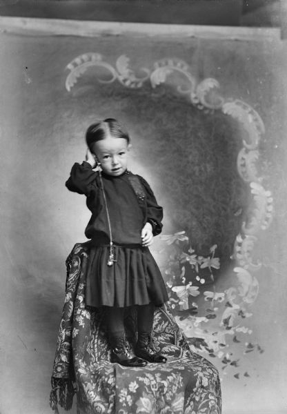A young child stands on a chair in a photography studio and holds a pocket watch to its ear in front of a painted backdrop.