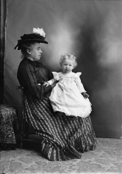 A woman wearing a hat, blouse, and long skirt sits in profile in a studio with a small girl wearing a white dress on her lap. They are in front of a studio backdrop.