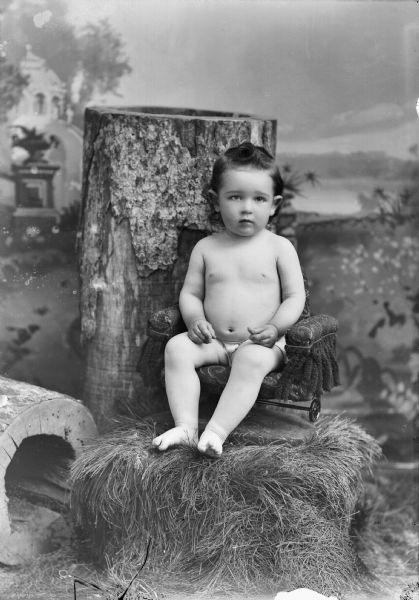A studio portrait of a young child wearing only shorts seated on a small chair which is on a bale of hay near two logs in front of a painted backdrop.
