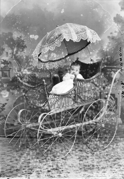 A studio portrait of an infant seated in a baby carriage under a lace-covered umbrella in front of a painted backdrop. "Moran Baby" written in negative.