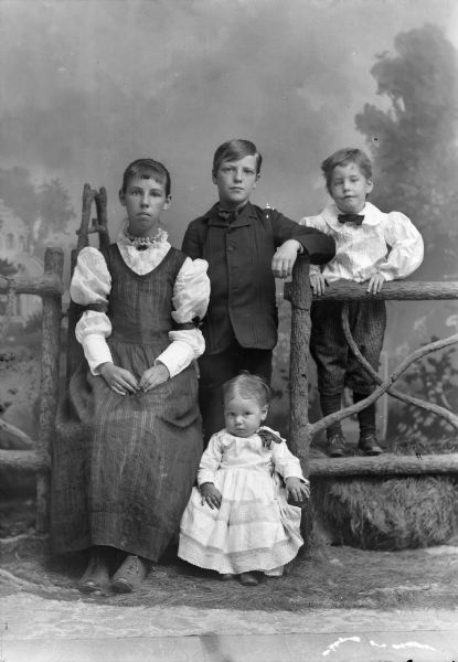 A studio portrait of group of children next to a wooden fence in front of a painted backdrop. The girl is sitting and wearing a lace-collared dress with ribbons tied on the sleeves near the elbow. The baby girl is in a dress and sitting low to the ground. The two boys are behind them, one in a suit jacket, the other in a puffy-sleeved shirt.