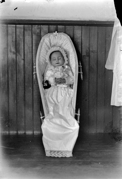 An infant in a bonnet holding a flower in an open coffin leaning upright against a wall.