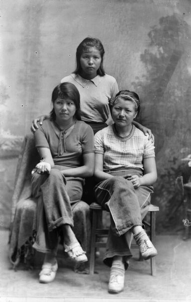 A studio portrait of three Native American women in front of a painted backdrop, one standing and two seated. The two women in front are wearing blouses and trousers.